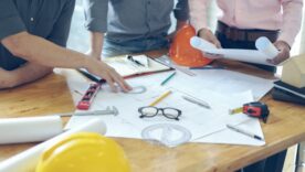 Three males in smart tops around a table with construction drawings and a construction hat