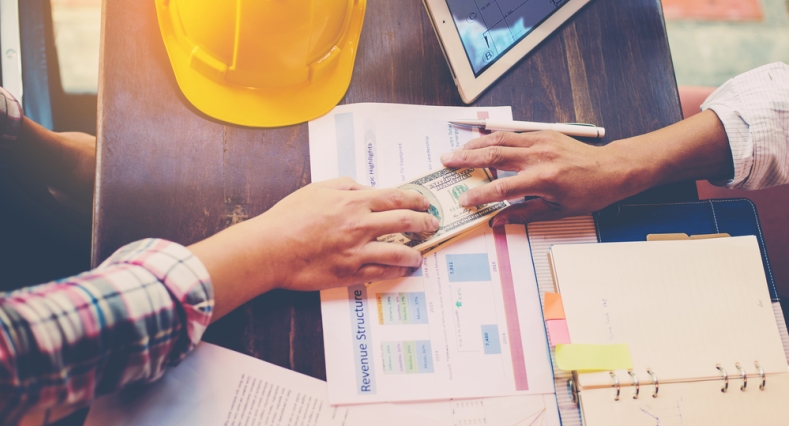 Wooden table with a construction hat and two individuals passing paper money over the top