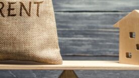 A wooden plank balancing equally with one side having a sack on with the word rent and the other side a wooden block house