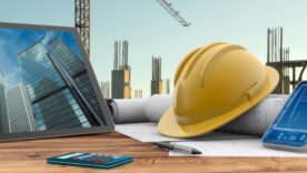 Tablet showing office buildings on a table filled with construction plans, a hard hat, calculator and documents. In the background is the start of the construction of the buildings