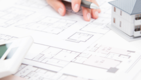 A person's hand holding a pen over an architectural drawing with a model house and a calculator in the background