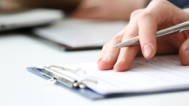 Hand of businessman in suit filling and signing with silver pen partnership agreement form clipped to pad closeup.