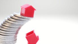 A red house on a slanting stack of coins. Below one house that already fell.