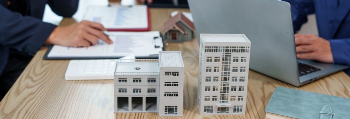 A tabletop with architectural models of buildings, a laptop, and documents, as two business professionals discuss real estate plans.