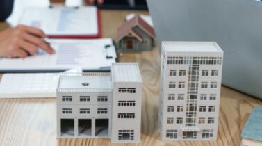 A tabletop with architectural models of buildings, a laptop, and documents, as two business professionals discuss real estate plans.