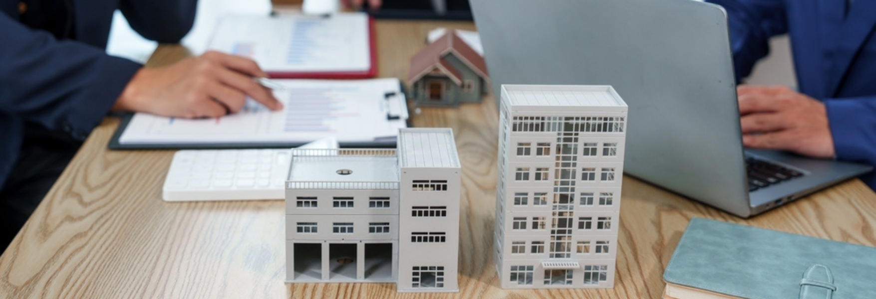 A tabletop with architectural models of buildings, a laptop, and documents, as two business professionals discuss real estate plans.