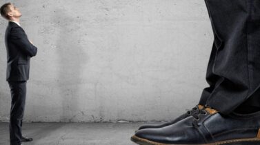 Tiny businessman looking up on huge legs of another businessman.