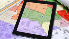 Tablet displaying a colour-coded property and zoning map on top of survey plans, representing land boundaries and ownership.