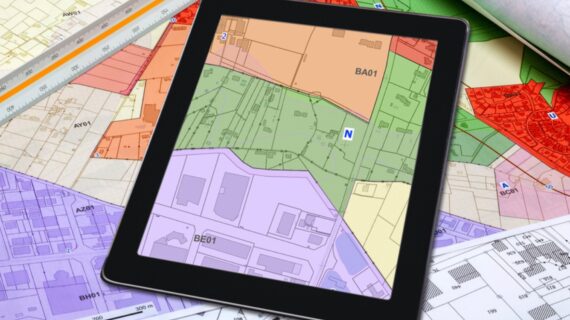 Tablet displaying a colour-coded property and zoning map on top of survey plans, representing land boundaries and ownership.