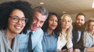 A diverse group of professionals sits in a modern conference room, focused intently during a meeting.