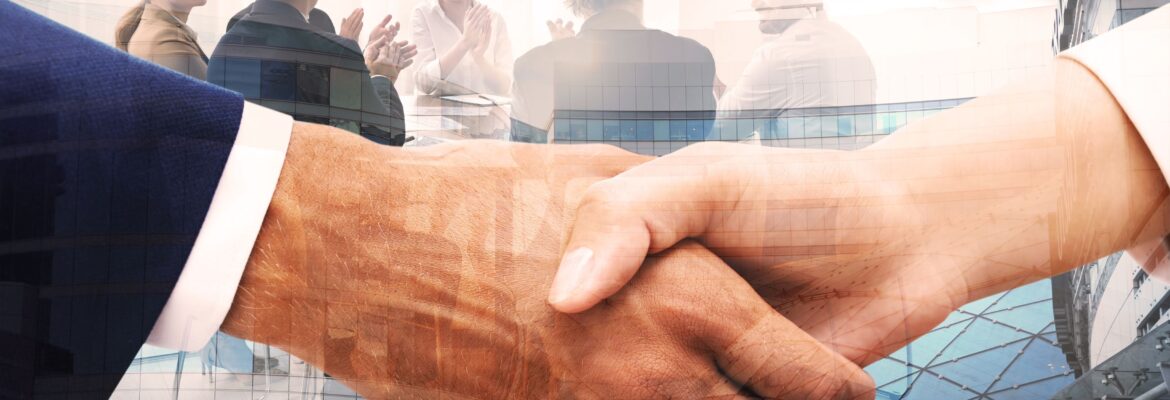 A close up of a handshake, in the background there are several business people sitting around a table