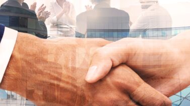 A close up of a handshake, in the background there are several business people sitting around a table