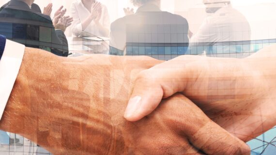 A close up of a handshake, in the background there are several business people sitting around a table