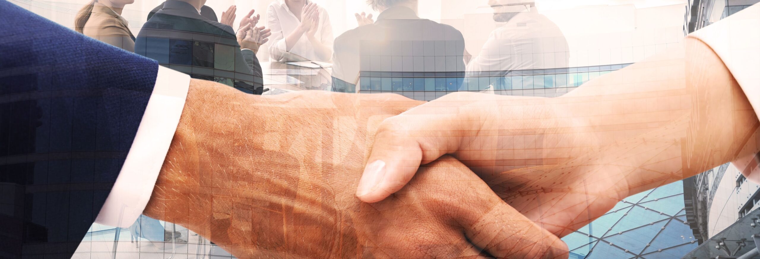 A close up of a handshake, in the background there are several business people sitting around a table