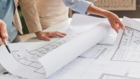 Two people are examining architectural blueprints on a table, one pointing with a pen as they discuss plans.