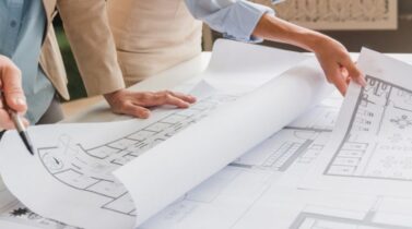 Two people are examining architectural blueprints on a table, one pointing with a pen as they discuss plans.