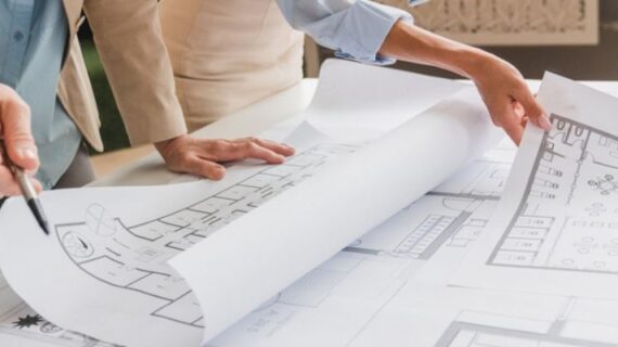 Two people are examining architectural blueprints on a table, one pointing with a pen as they discuss plans.