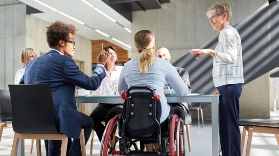 A diverse group of professionals engaged in a meeting, with one person standing and another in a wheelchair participating.