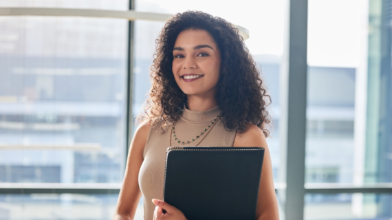 Woman with curly hair holding a black folder, standing in front of large office windows with a cityscape background.