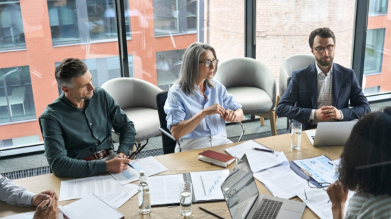 Business team in a meeting room with documents, laptops, and glasses of water, discussing work at a conference table near large windows.
