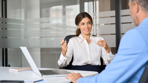 Two professionals having a discussion in a modern office, with a laptop open on the table between them.