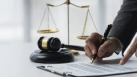 Person signing a legal document with a gavel and scales of justice on the desk, symbolizing law and legal agreements.
