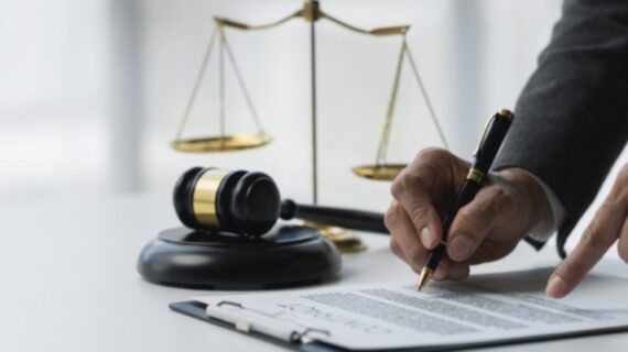 Person signing a legal document with a gavel and scales of justice on the desk, symbolizing law and legal agreements.