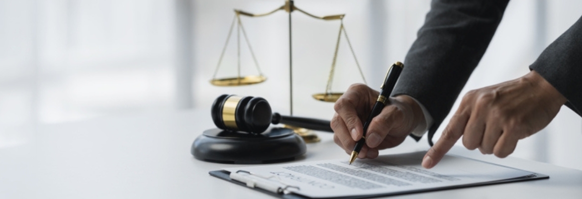 Person signing a legal document with a gavel and scales of justice on the desk, symbolizing law and legal agreements.