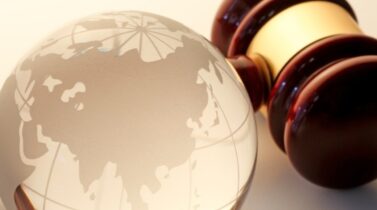 A wooden judge's gavel beside a transparent globe etched with a world map, symbolizing international law and justice.