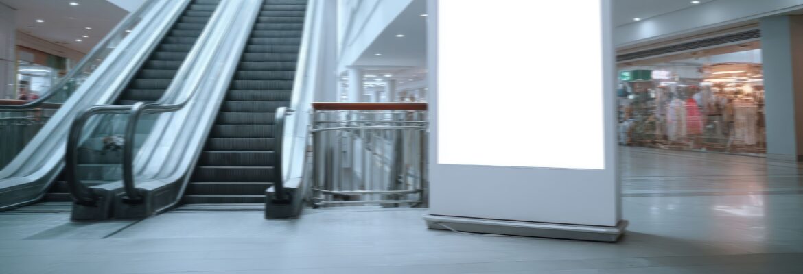 A blank billboard sits next to escalators inside a commercial building or shopping center, ready for advertising