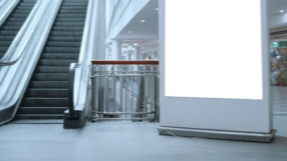 A blank billboard sits next to escalators inside a commercial building or shopping center, ready for advertising