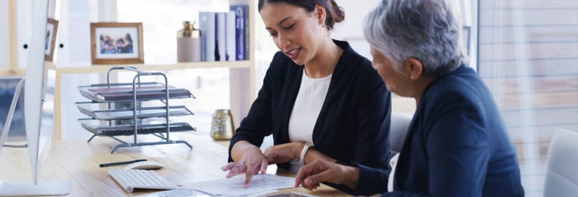 Two businesswomen reviewing documents together at an office desk with a computer and document trays nearby.