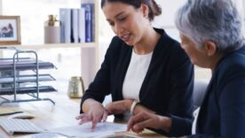 Two businesswomen reviewing documents together at an office desk with a computer and document trays nearby.