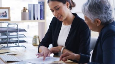 Two businesswomen reviewing documents together at an office desk with a computer and document trays nearby.