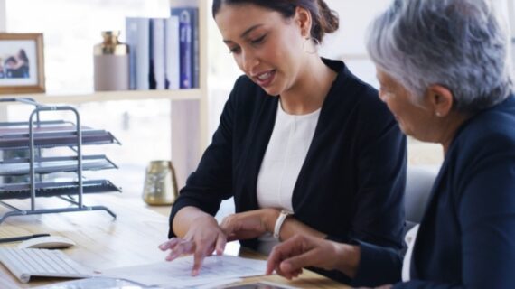 Two businesswomen reviewing documents together at an office desk with a computer and document trays nearby.