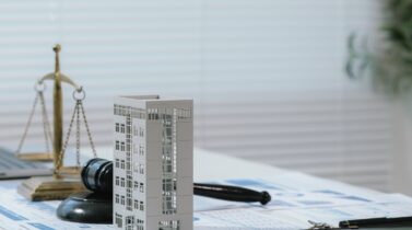 Justice scale and judge gavel beside building model and business reports, representing real estate law, fair property transactions, and housing regulations