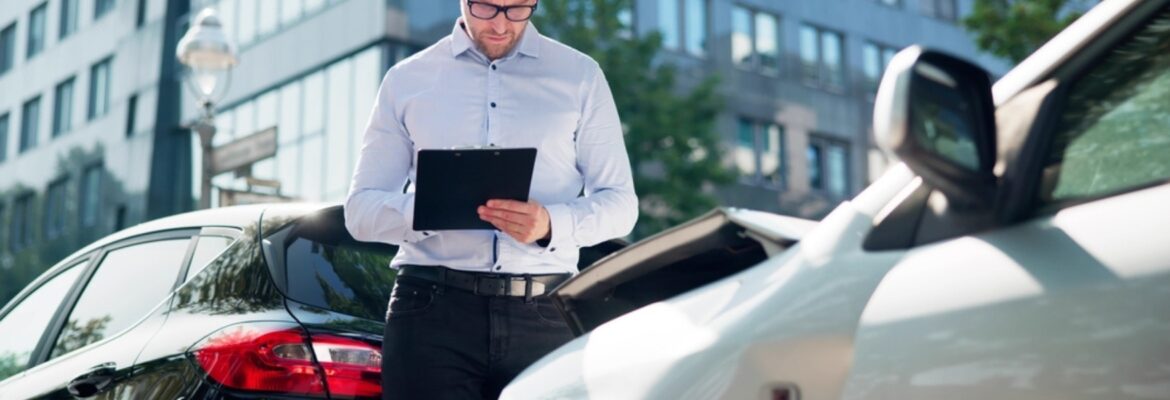 Man in glasses inspects and takes notes on a clipboard near two cars after a minor collision.