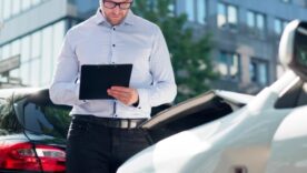 Man in glasses inspects and takes notes on a clipboard near two cars after a minor collision.