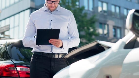 Man in glasses inspects and takes notes on a clipboard near two cars after a minor collision.