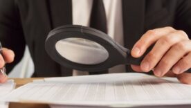 Person in a suit using a magnifying glass to closely examine documents on a desk, holding a pen in the other hand.