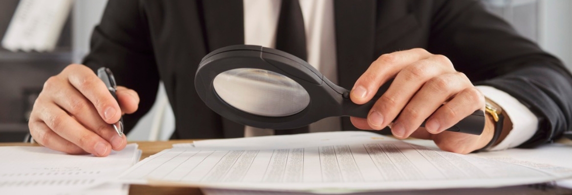 Person in a suit using a magnifying glass to closely examine documents on a desk, holding a pen in the other hand.