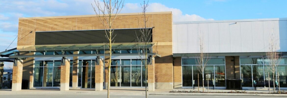 Modern brick-and-metal commercial building with large glass storefronts, covered entrance, leafless trees, and blue sky.