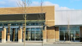 Modern brick-and-metal commercial building with large glass storefronts, covered entrance, leafless trees, and blue sky.