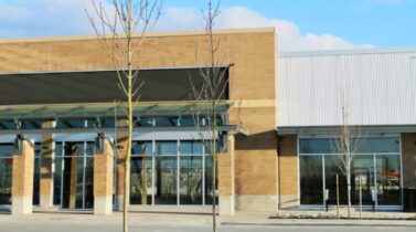 Modern brick-and-metal commercial building with large glass storefronts, covered entrance, leafless trees, and blue sky.