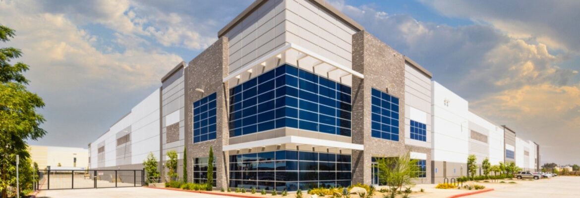 Modern industrial warehouse with blue glass-fronted entrance, stone accents, gated driveway, landscaping, and partly cloudy sky.