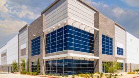 Modern industrial warehouse with blue glass-fronted entrance, stone accents, gated driveway, landscaping, and partly cloudy sky.