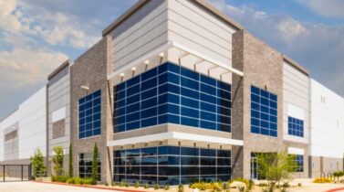 Modern industrial warehouse with blue glass-fronted entrance, stone accents, gated driveway, landscaping, and partly cloudy sky.