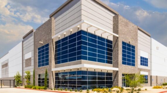 Modern industrial warehouse with blue glass-fronted entrance, stone accents, gated driveway, landscaping, and partly cloudy sky.