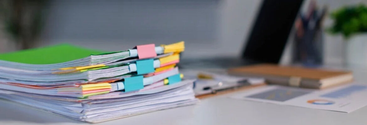 Stack of colorful, binder-clipped documents on a desk beside a laptop, notebook, and stationery.