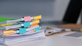 Stack of colorful, binder-clipped documents on a desk beside a laptop, notebook, and stationery.